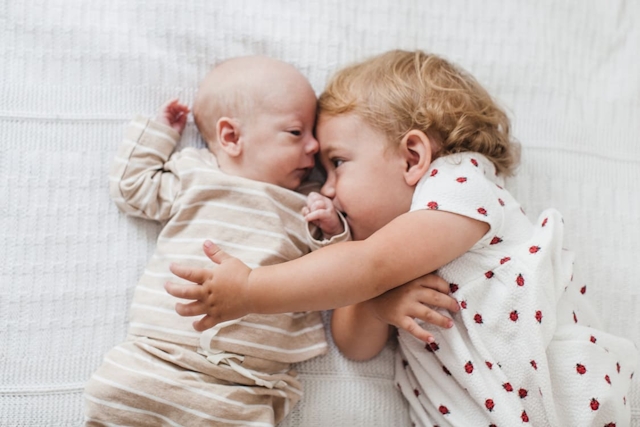 Schwester liegt mit neugeborenem Bruder auf dem Bett und umarmt ihn