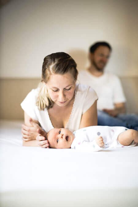 Mutter liegt bäuchlings auf dem Bett und beugt sich über ihr Baby. der Vater sitzt im Hintergrund.