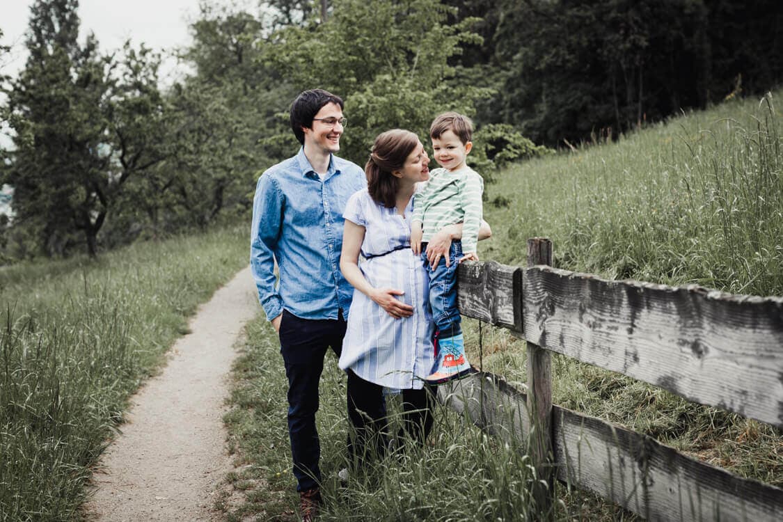 Junge sitzt auf einem Zaun. Die schwangere Mutter hält ihn fest.