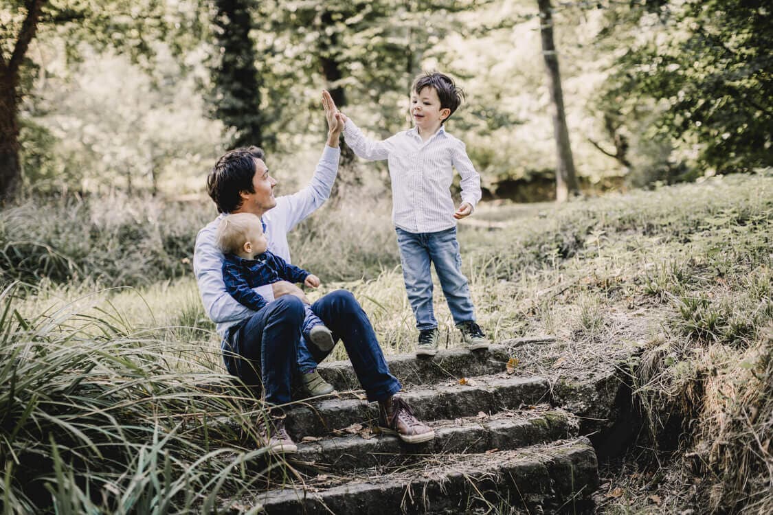 Vater sitzt mit Kind auf dem Schoß auf einer Treppe und gibt dem anderen Sohn ein high five
