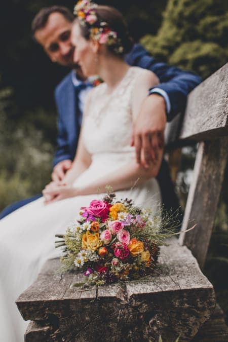 Brautstrauß auf einer Bank. Brautpaar im Hintergrund.