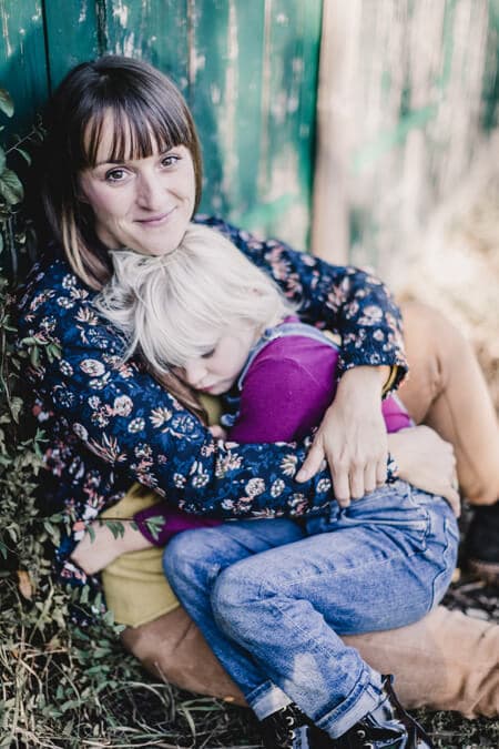 Frau lehnt an einer Scheune und hat ihre Tochter auf dem Schoß