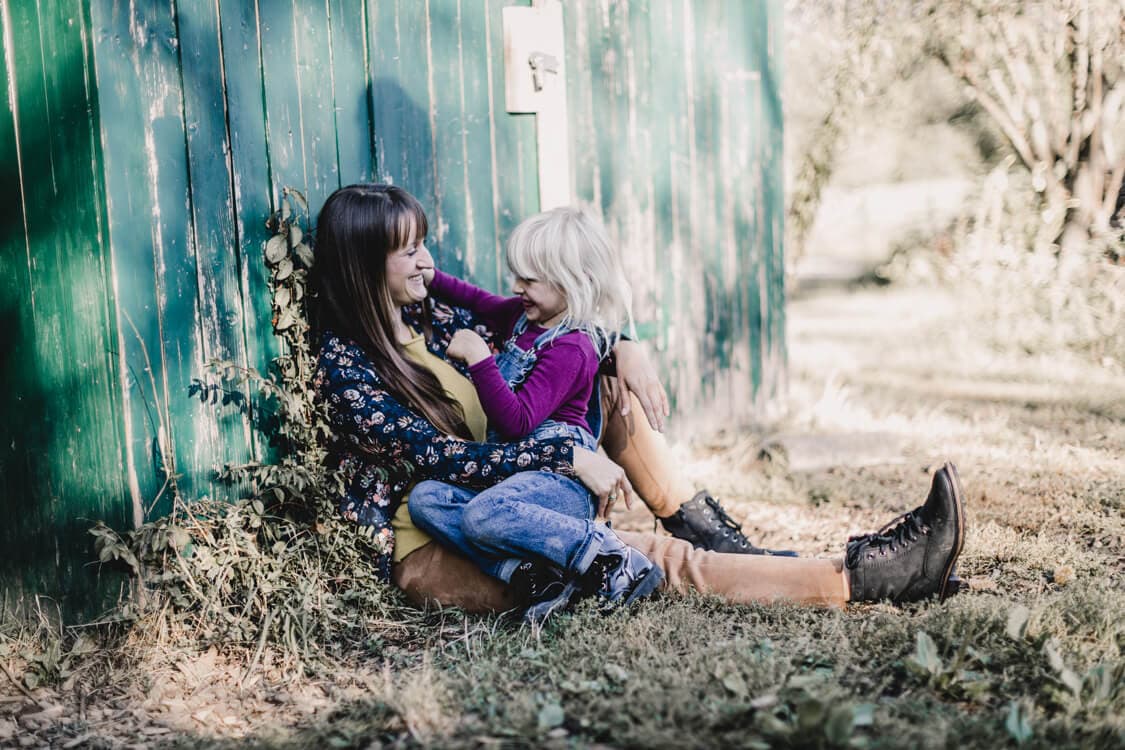 Frau lehnt an einer Scheune und hat ihre Tochter auf dem Schoß und albert mit ihr herum