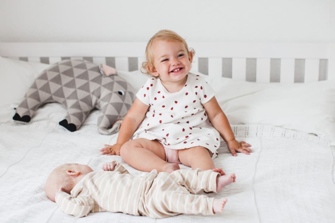 Mädchen sitzt lachend auf dem Bett. Vor ihr liegt ihr neugeborener Bruder.