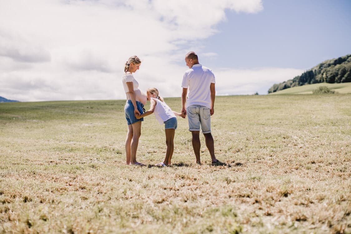 Familie Hand in Hand auf einem Feld. Tochter küsst den Babybauch der Mutter.