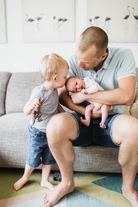 Papa mit Baby auf dem Arm. Großer Bruder gibt dem Baby einen Kuss.