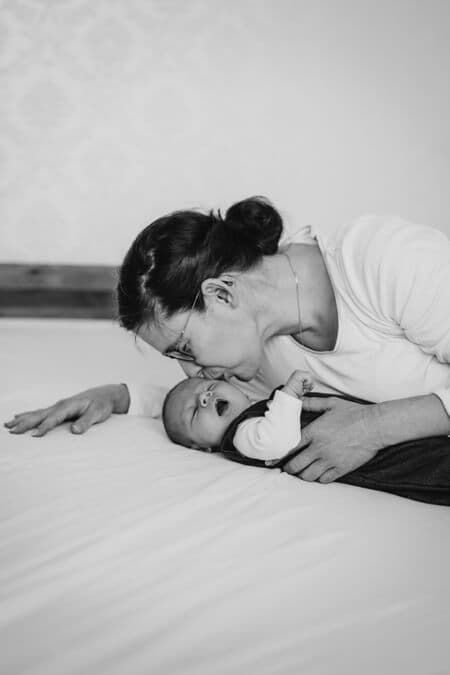 Baby liegt auf dem Bett und die Mutter beugt sich zu ihm um es zu küssen