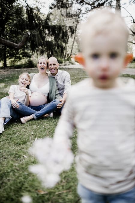 Kleiner Junge läuft auf die Kamera zu. Familie lacht im Hintergrung.