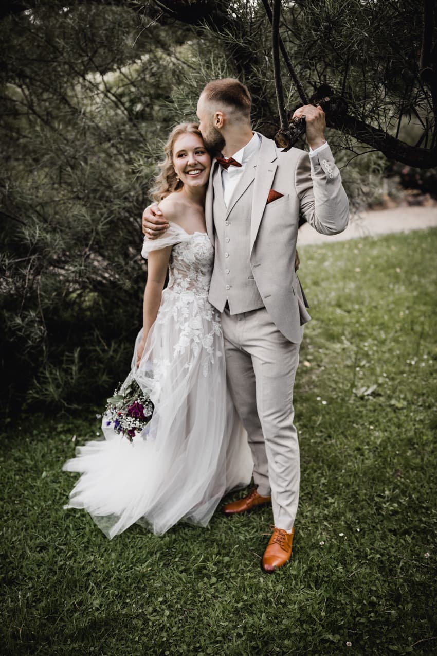 Brautpaar unter Baum, Bräutigam küsst Braut auf Kopf romantisches Hochzeitsfoto