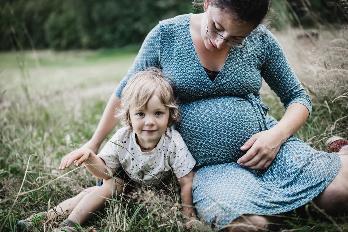 Junge sitzt neben seiner schwangeren Mama auf einer Wiese