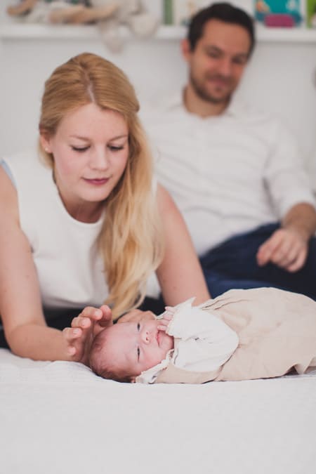 Mama beugt sich zu ihrem Neugeborenen vor. Papa sitzt im Hintergrund.