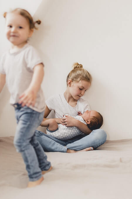 Mädchen hält ihren Bruder im Arm. Kleiner Schwester hüpft im Vordergrund.