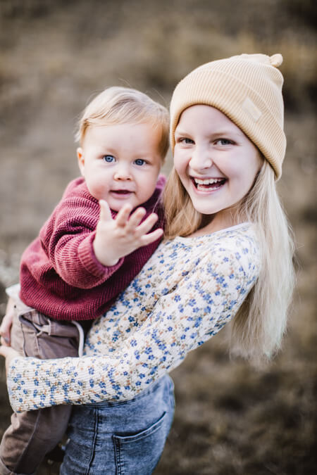 Schulkind mit kleinem Bruder auf dem Arm. Der Junge Winkt in die Kamera.