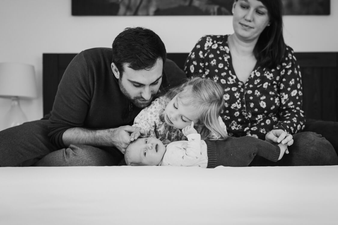 Baby liegt auf dem Bett. Vater und Schwester beugen sich zu ihm. Mutter sitzt im Hintergrund.