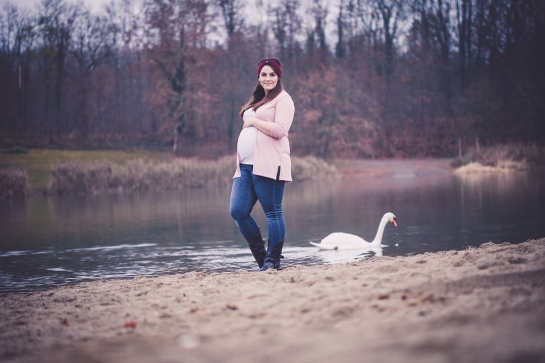 Schwangere Frau steht am See. Im Hintergrund schwimmt ein Schwan.