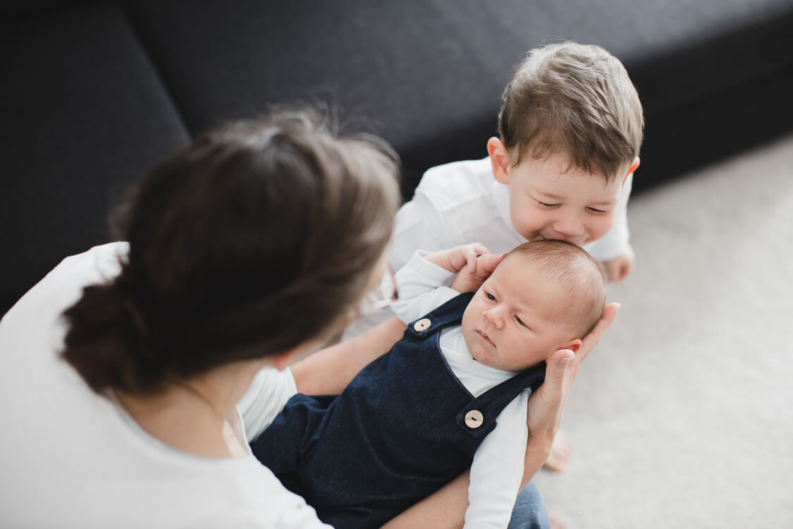 Mutter hält ihr Neugeborenes vor sich. Der große Sohn gibt dem Baby einen Kuss auf den Kopf.