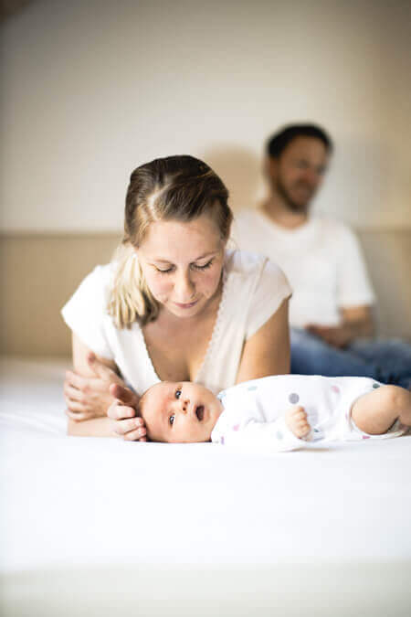 Mutter liegt bäuchlings auf dem Bett und beugt sich über ihr Baby. der Vater sitzt im Hintergrund.