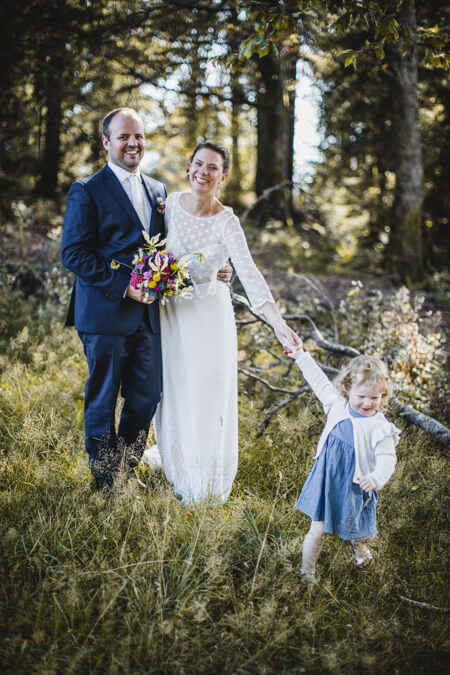 Brautpaar steht an einer Waldlichtung und Tochter zieht die Mutter am Arm