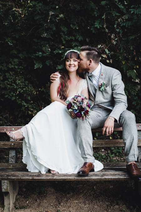Brautpaar sitzt auf der Lehne einer Bank. Der Mann küsst seine Frau.