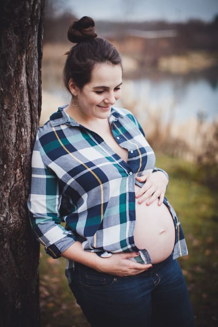 Frau mit Babybauch und Flanellhemd lehnt an einem Baum