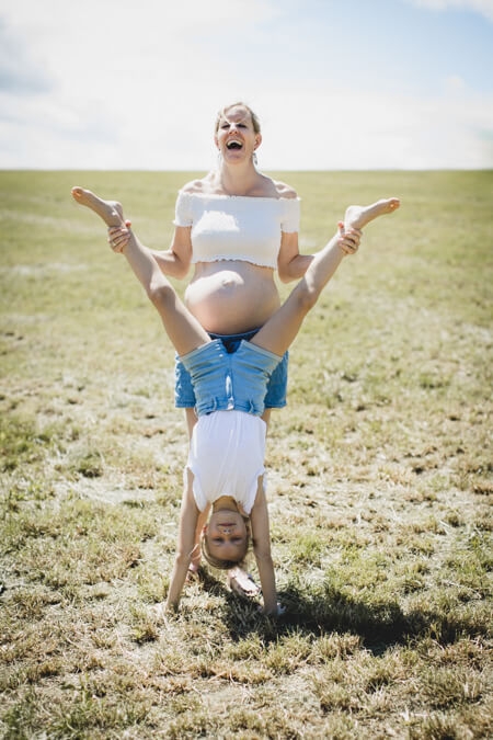 Frau mit Babybauch steht auf einer Wiese und lacht. Sie hält die Beine ihrer Tochter die vor ihr einen Handstand macht.