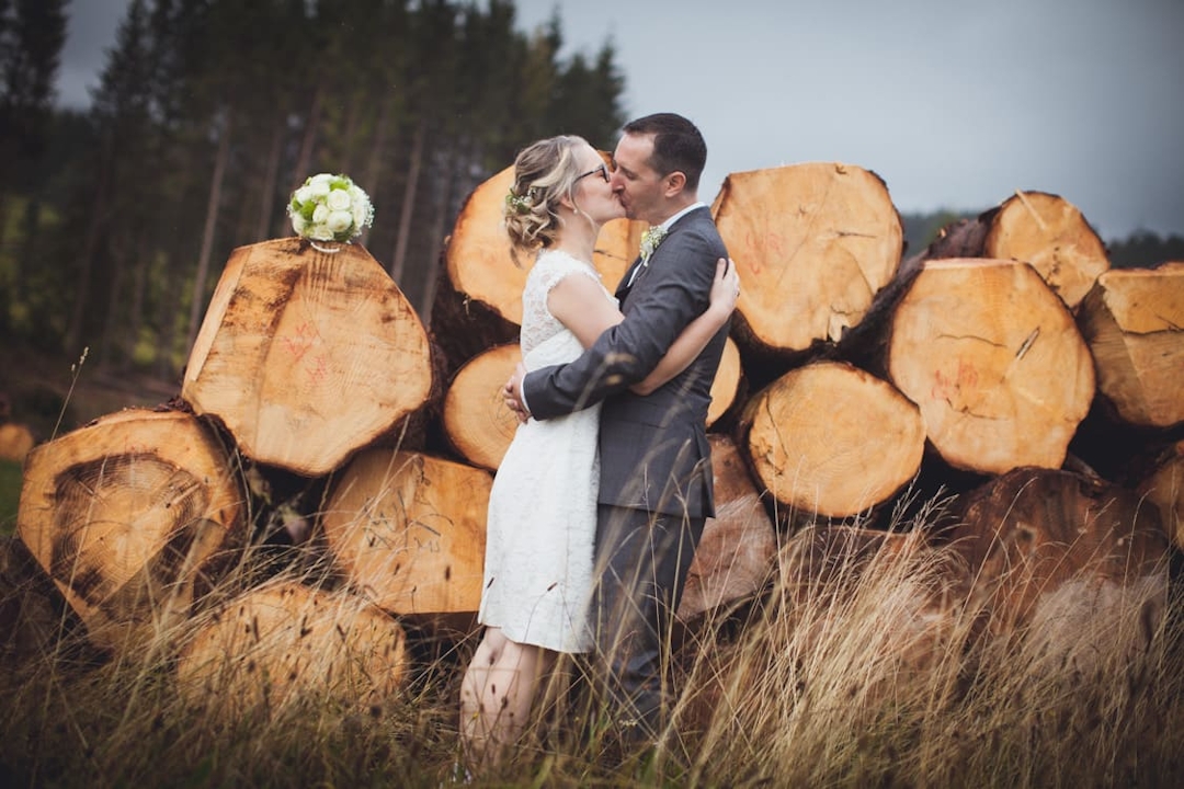 Brautpaar küsst sich vor einem Stapel von Baumstämmen