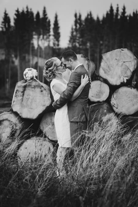 SW Foto von küssendem Brautpaar vor einem Stapel Baumstämmen