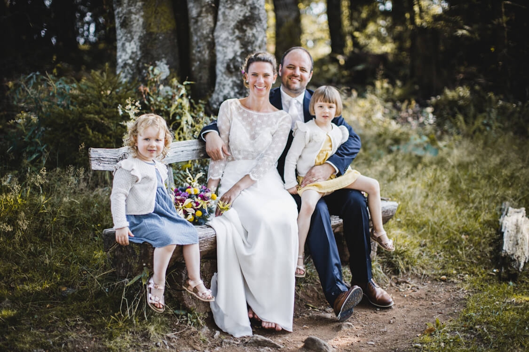 Braut und Bräutigam sitzen mit ihren beiden Töchtern auf einer Bank im Wald