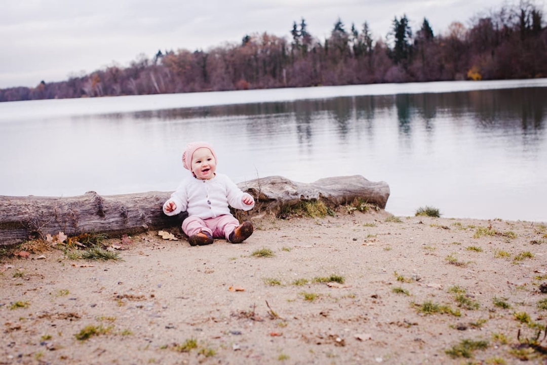 Kleines Mädchen sitzt vor einem liegenden Baumstamm. Im Hintergrund ist ein See.