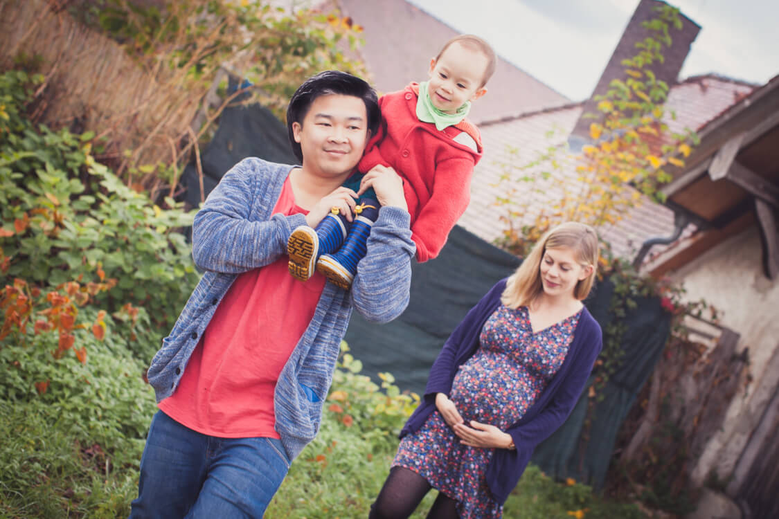 Kleines Kind sitzt auf der Schulter von seinem Papa. Frau mit Babybauch im Hintergrund.