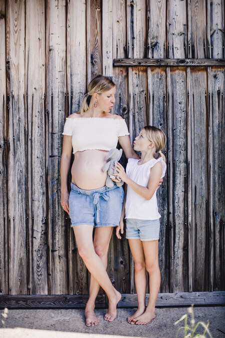 frau mit Babybauch und ihrer Tochter lehnen an einem Scheunentor. Die Tochter hat ein Kuscheltier in der Hand.